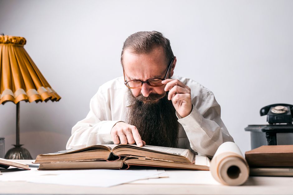 Bearded man with eyeglasses focused on reading a large book at a vintage desk.